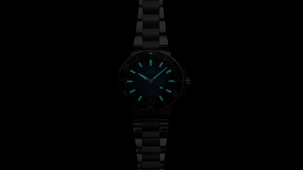 Oris Aquis watch with a striking blue dial, stainless steel case, and a unidirectional rotating bezel, showcasing its sleek design and robust functionality. Glow in the dark view.