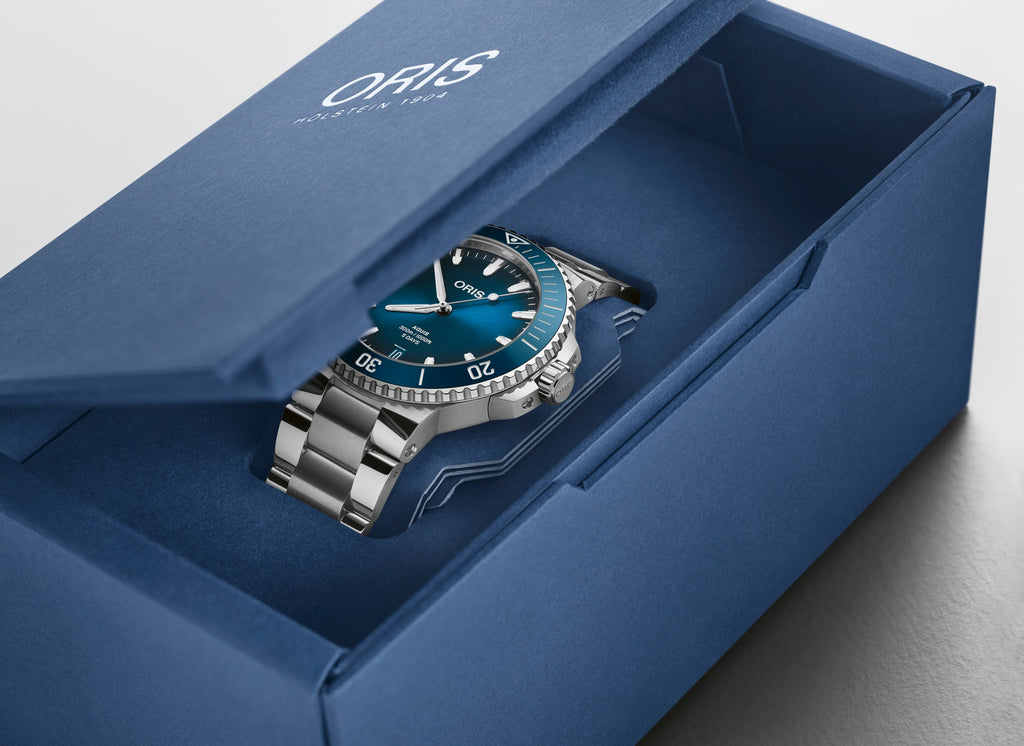 Oris Aquis watch with a striking blue dial, stainless steel case, and a unidirectional rotating bezel, showcasing its sleek design and robust functionality. In sustainable box view.