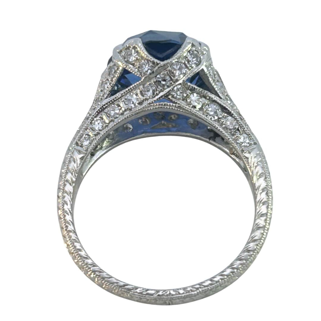 Edwardian-inspired platinum ring with a 6.16ct round blue sapphire and 54 diamond accents, detailed with milgrain and engraving.