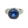 Edwardian-inspired platinum ring with a 6.16ct round blue sapphire and 54 diamond accents, detailed with milgrain and engraving.