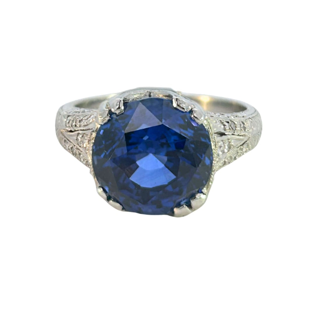 Edwardian-inspired platinum ring with a 6.16ct round blue sapphire and 54 diamond accents, detailed with milgrain and engraving.