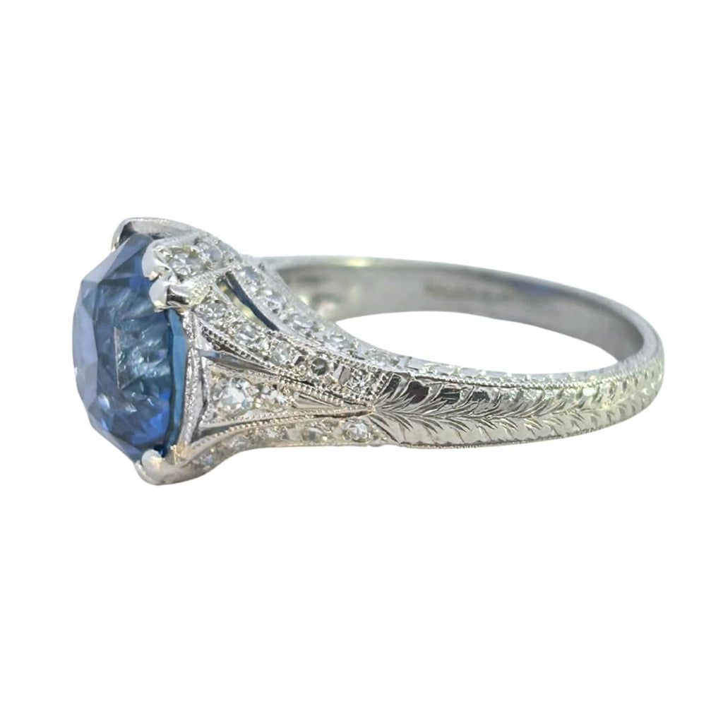 Edwardian-inspired platinum ring with a 6.16ct round blue sapphire and 54 diamond accents, detailed with milgrain and engraving.